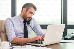 Hombre maduro confiado en camisa y corbata trabajando en la computadora portátil y hablando por teléfono mientras está sentado en su lugar de trabajo