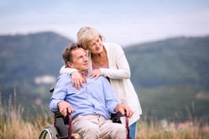 Senior woman pushing man sitting in wheelchair oustide in green autumn nature, hugging