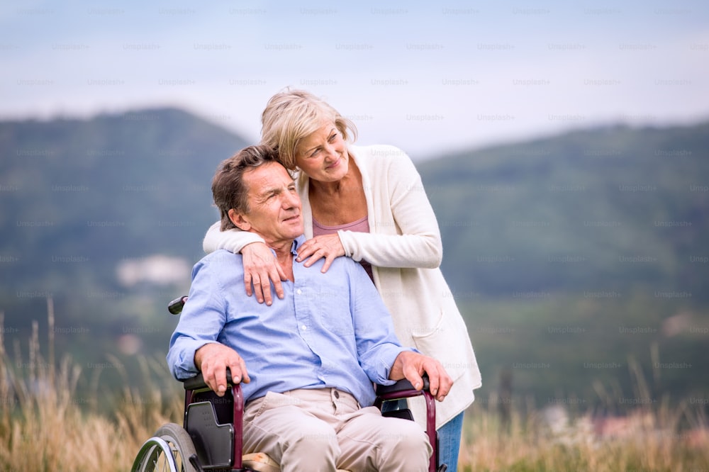 Ältere Frau schiebt Mann sitzt im Rollstuhl Oustide in grüner Herbstnatur, umarmt