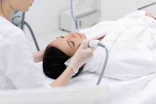 Une jeune femme détendue reçoit un massage facial en cavitation par cosmétologue. Elle est allongée sur la table. Ses yeux sont fermés de jouissance