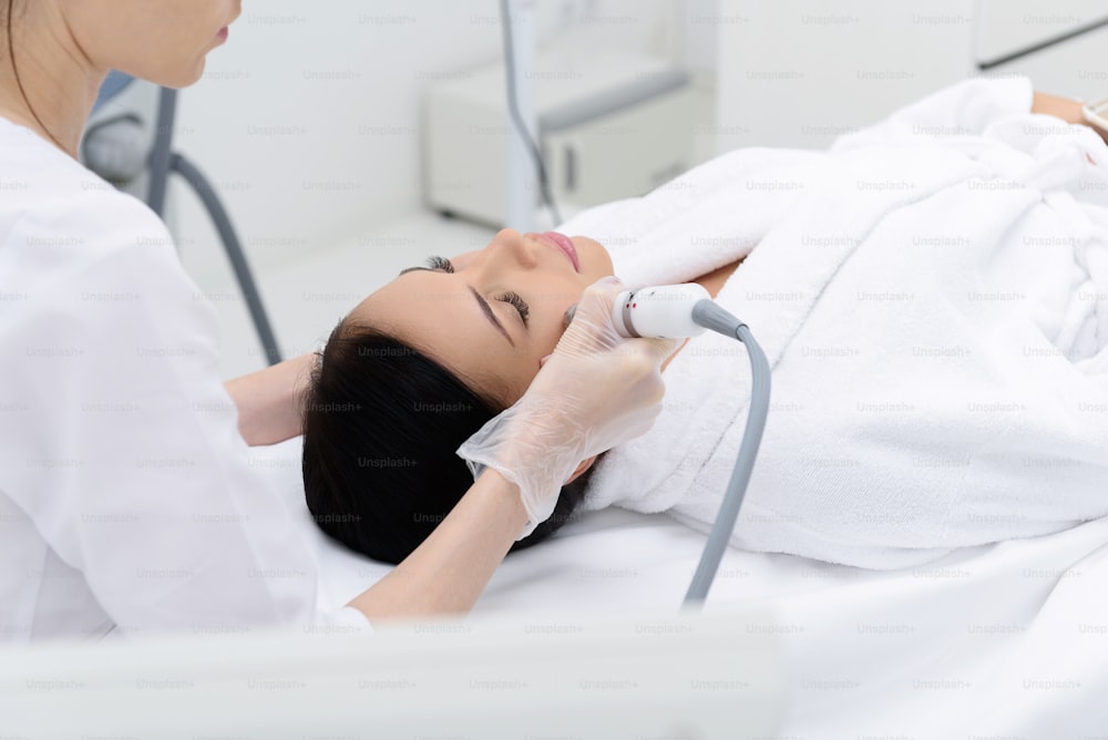Una mujer joven relajada está recibiendo un masaje facial de cavitación por parte de un cosmetólogo. Está acostada sobre la mesa. Sus ojos están cerrados de placer