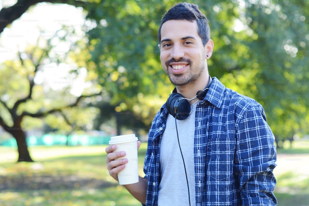 Portrait of young handsome man with headphones and drinking coffee in a park. Outdoors.