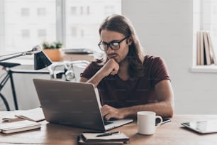 Jeune homme réfléchi aux cheveux longs regardant l’ordinateur portable et tenant la main sur le menton tout en étant assis à son lieu de travail au bureau