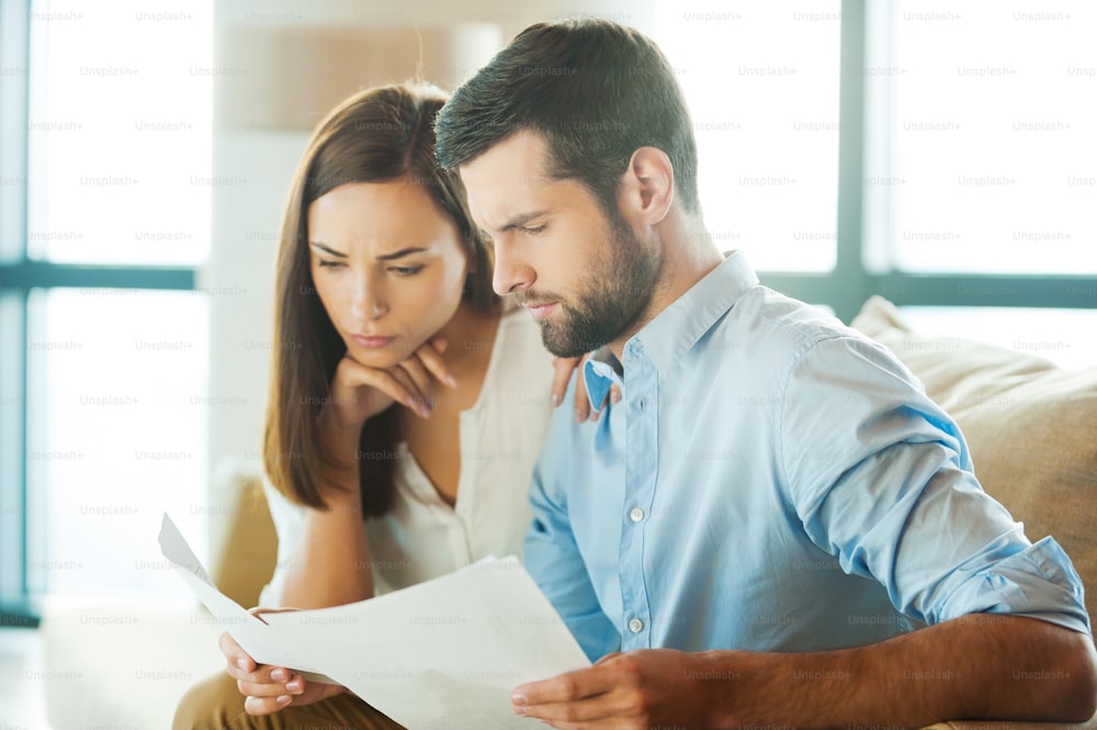 Concentrated young man holding documents and looking at them while woman sitting close to him and holding hand on chin