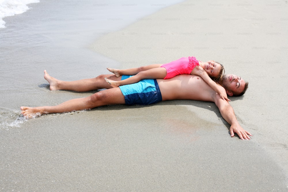 Pai deitado na areia com a filha na praia. Token de fotos de cima.