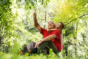 Grandfather pointing with finger his granddaughter something up in forest.