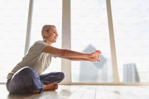 Una giovane donna felice sta facendo yoga con divertimento a casa. È seduta vicino alla finestra e allunga le braccia in avanti. La signora sta ridendo