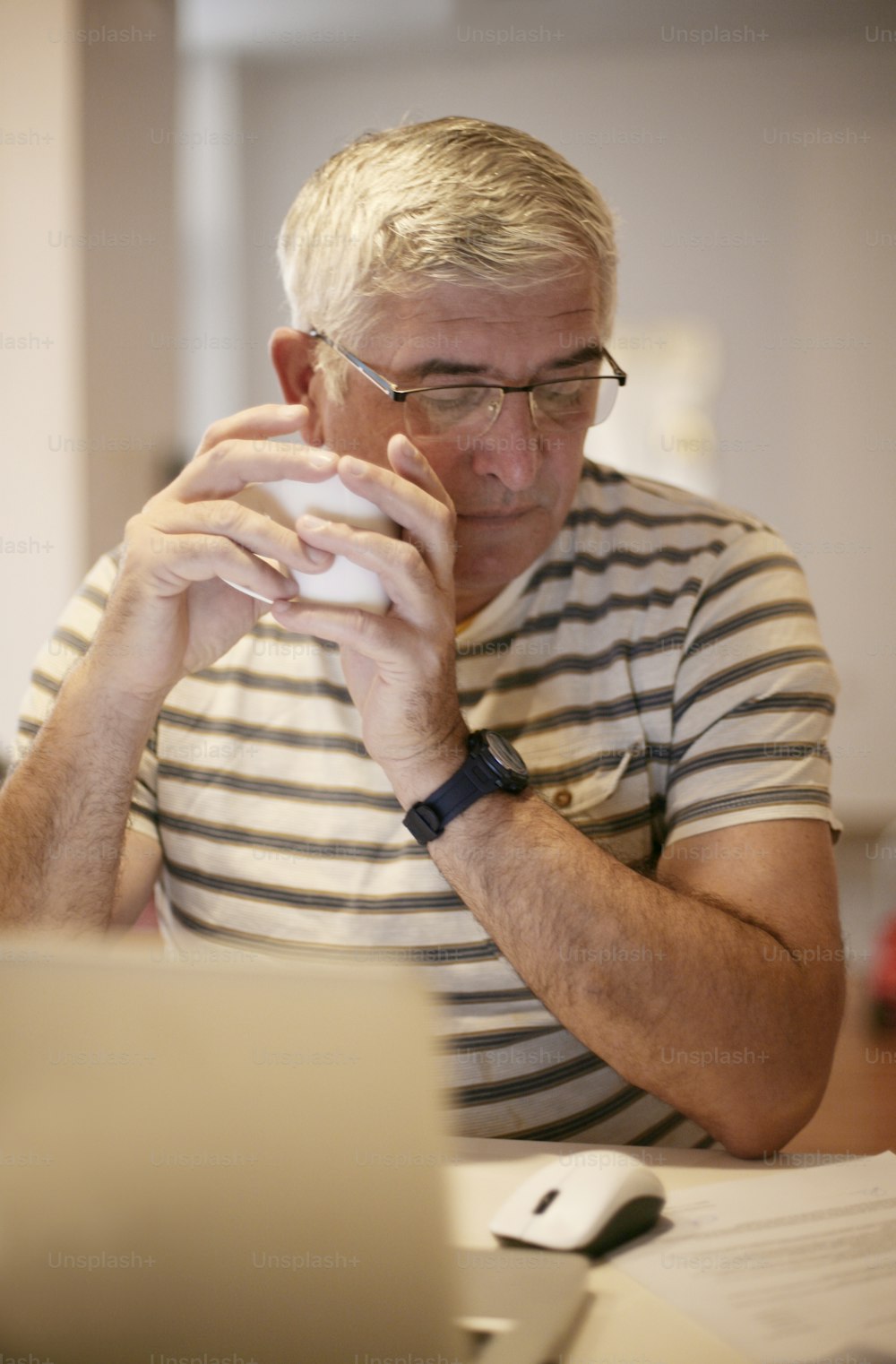 Uomo anziano che beve caffè e lavora sul laptop a casa.