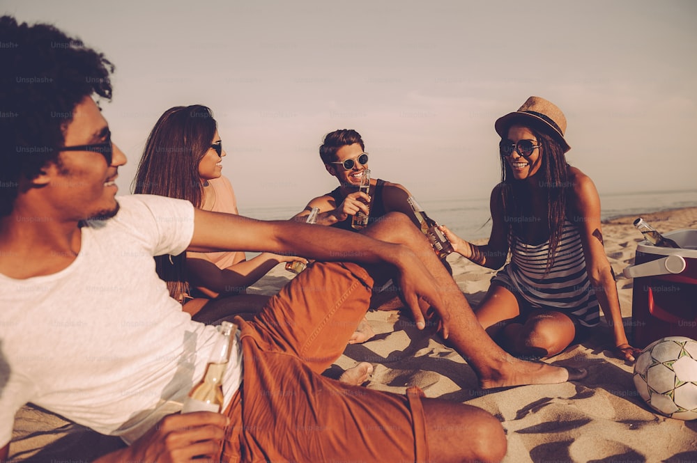 Giovani allegri che trascorrono del bel tempo insieme seduti sulla spiaggia e bevono birra