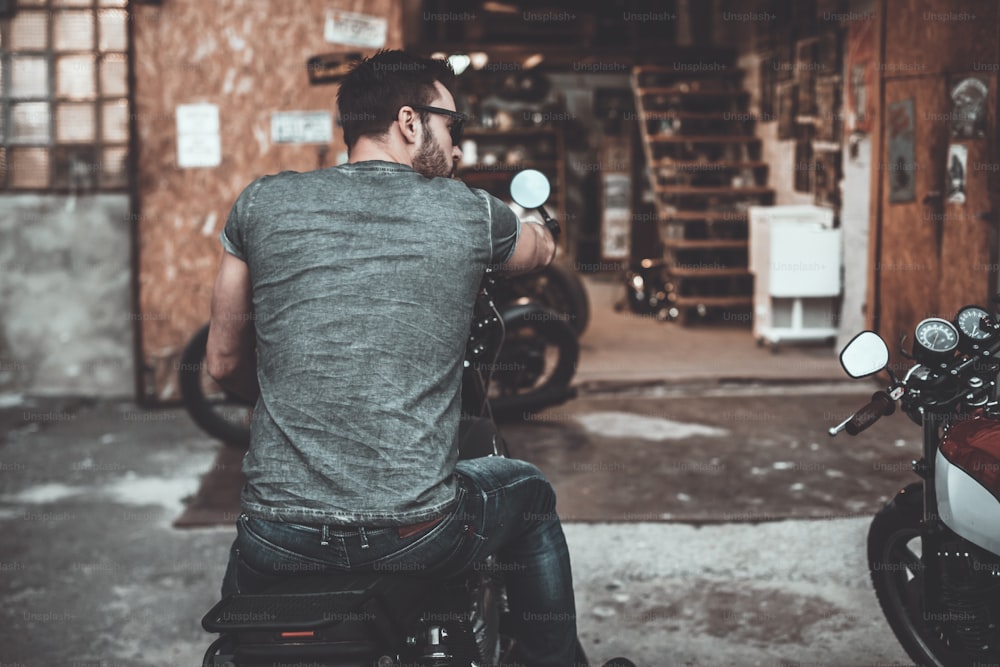 Rear view of confident young man sitting on his bike with motorcycle garage in the background