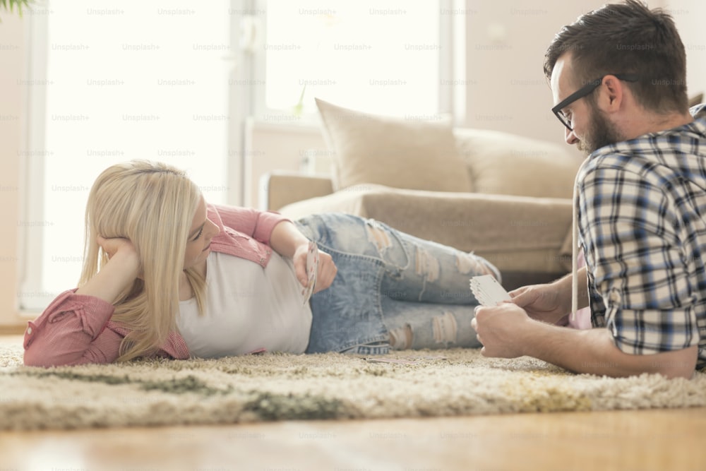 Couple in love lying on the carpet on the floor, playing cards and enjoying their leisure time. Selective focus, lens flare effect on the window.