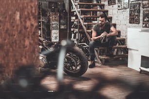 Selbstbewusster junger Mann, der Lappen hält und das Motorrad betrachtet, während er in der Werkstatt in der Nähe sitzt