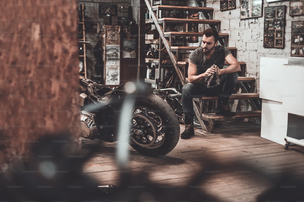Joven confiado sosteniendo un trapo y mirando la motocicleta mientras está sentado cerca de ella en el taller de reparación