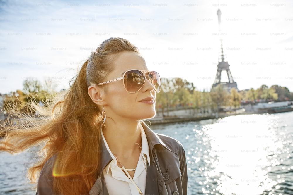 Escapadinhas de outono em Paris. Retrato da mulher elegante jovem em óculos de sol no aterro em Paris, France olhando para a distância