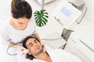 Un abile cosmetologo sta pulendo il viso maschile con apparecchiature per la cavitazione. L'uomo sta mentendo e si sta rilassando. I suoi occhi sono chiusi dal piacere