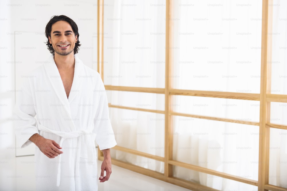 Good morning. Happy young man is standing in bathrobe at home. He is looking at camera and smiling