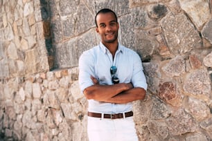 スマートカジュアルな服を着たハンサムな若いアフリカ人男性が腕を組んで、屋外の石の壁に立って笑顔でカメラを見ている