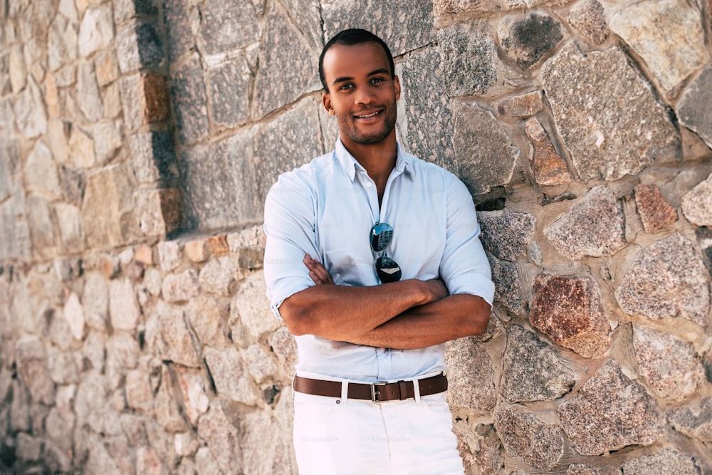 スマートカジュアルな服を着たハンサムな若いアフリカ人男性が腕を組んで、屋外の石の壁に立って笑顔でカメラを見ている