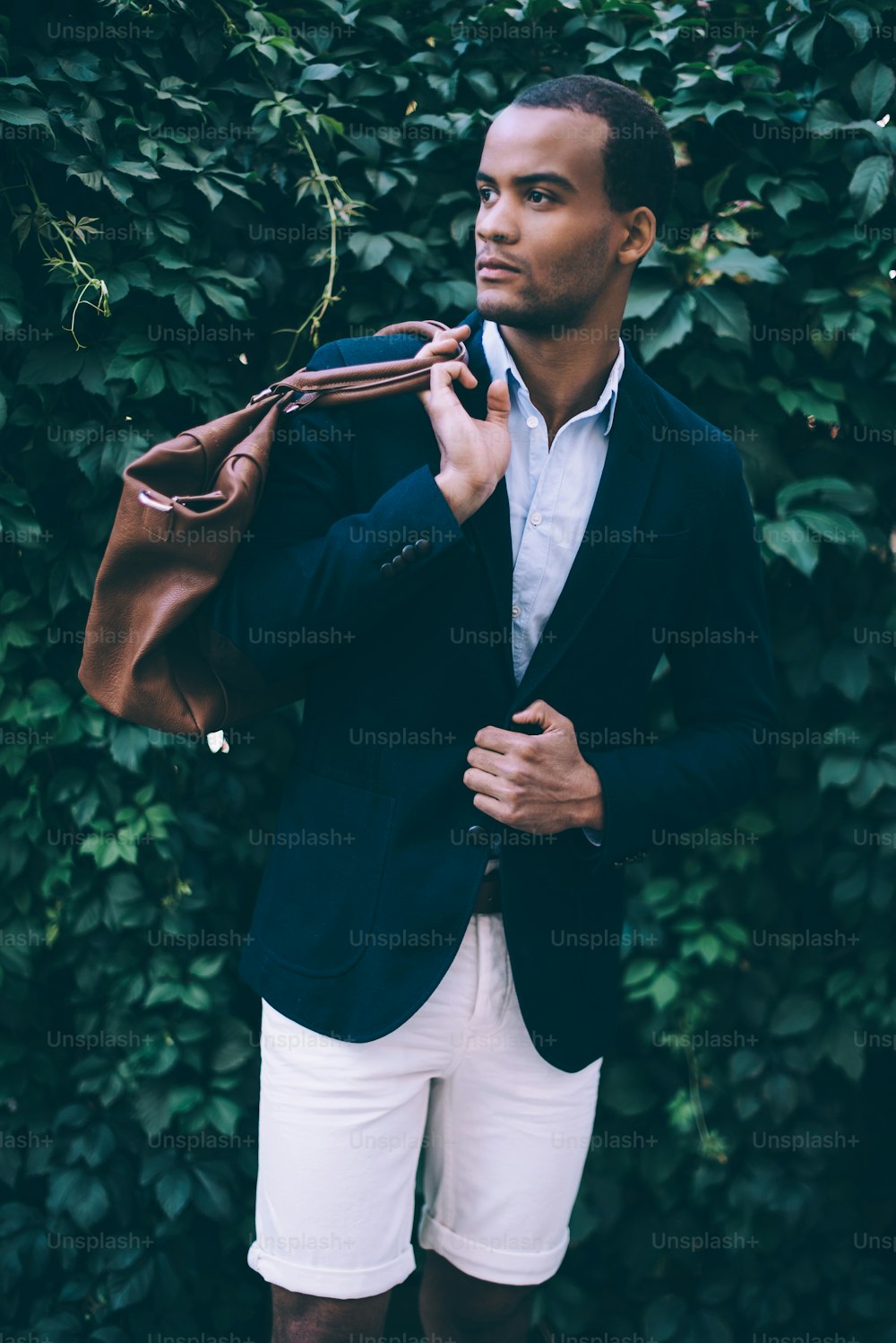 スマートカジュアルウェアのハンサムな若いアフリカの男性は、肩にバッグを運び、屋外の緑の植物の背景に立って見ています