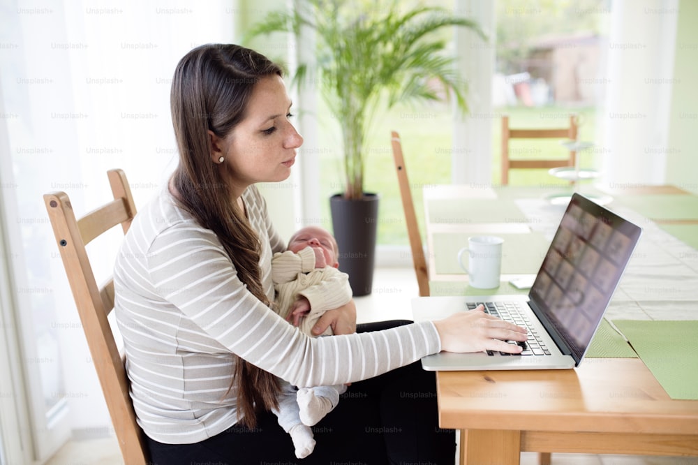 Beautiful young mother at home holding her newborn baby son in her lap, sitting at the table, writing on notebook keyboard