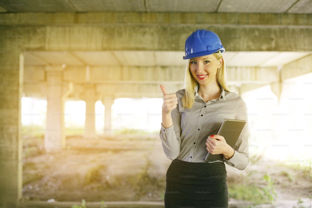 Blonde female architects holding digital tablet in arms showing ok with finger and looking at the camera.