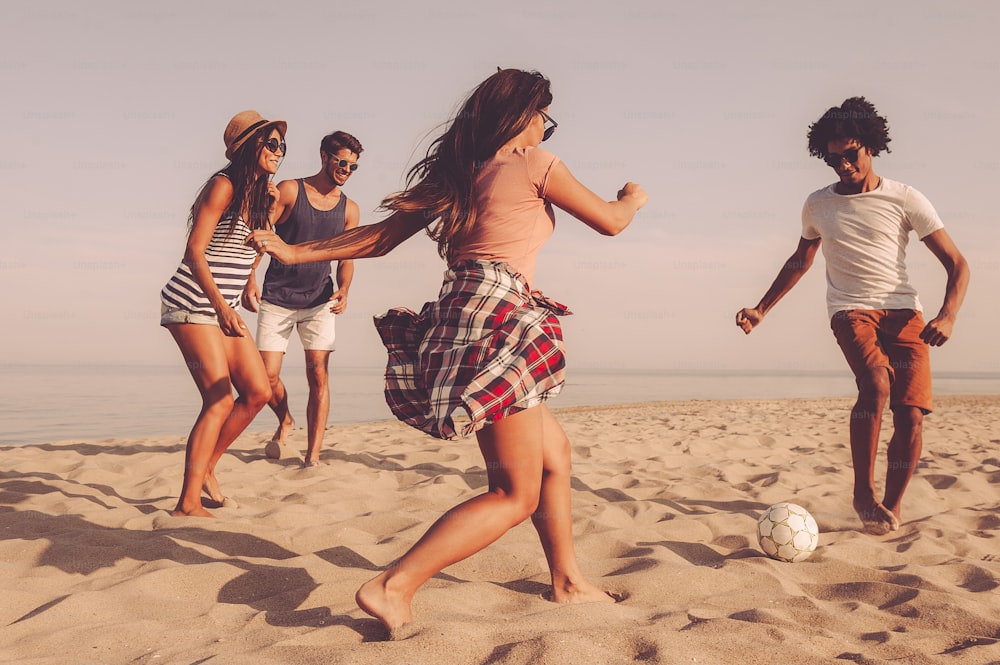 Gruppe fröhlicher junger Leute, die mit Fußball am Strand spielen, mit Meer im Hintergrund
