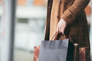 茶色の革のジャケットまたはトップコートを着たエレガントでモダンな女性が街の通りに立ち、買い物袋の束を持っています。女性の手の接写。ライフスタイル消費主義のテーマ。