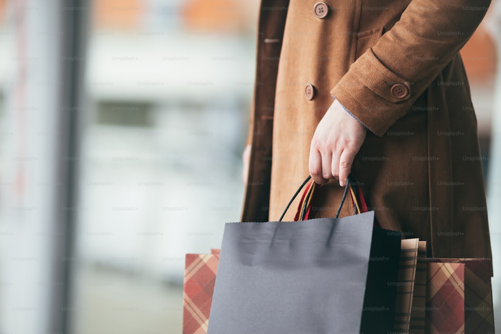 갈색 가죽 재킷이나 탑코트를 입은 우아하고 현대적인 여성이 도시 거리에 서서 쇼핑백을 잔뜩 들고 있다. 여자 손의 클로즈업 샷. 라이프 스타일 소비주의 테마.