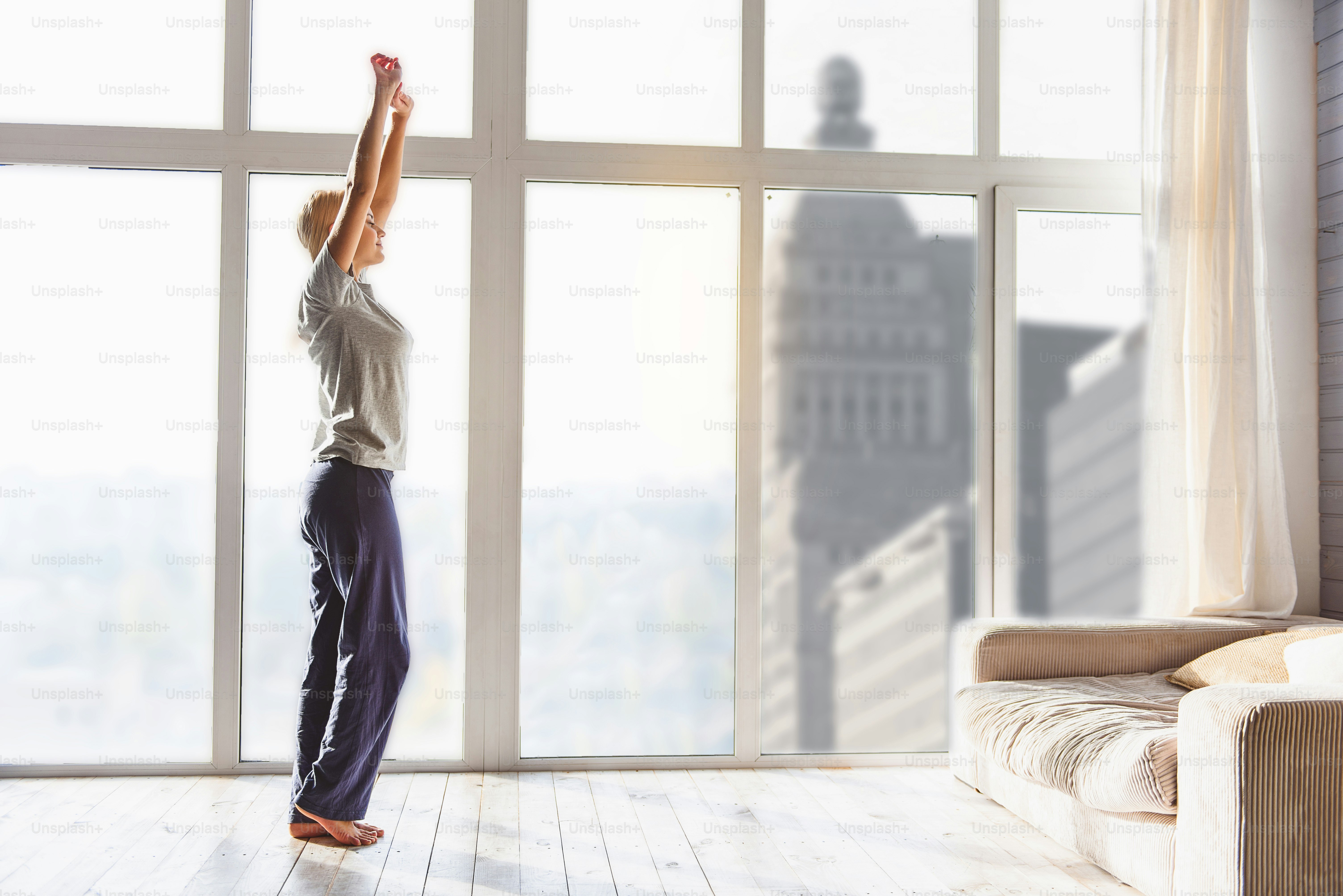 Schläfriges junges Mädchen macht morgens Sport. Sie steht in der Nähe des Fensters und streckt die Hände nach oben. Ihre Augen sind harmonisch geschlossen