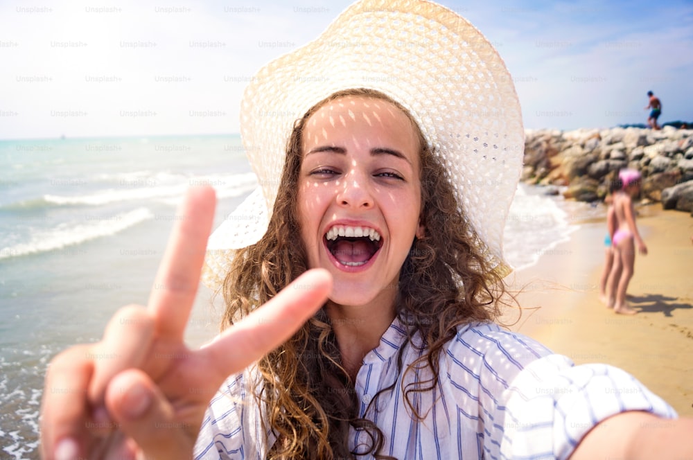 Beautiful young woman on beach wearing striped blue-and-white shirt and hat, laughing, taking selfie, showing peace sign. Enjoying time at seaside.