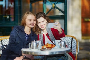 Duas meninas em um café ao ar livre parisiense, tomando café com croissant e tirando selfie usando o telefone celular. Conceito de amizade