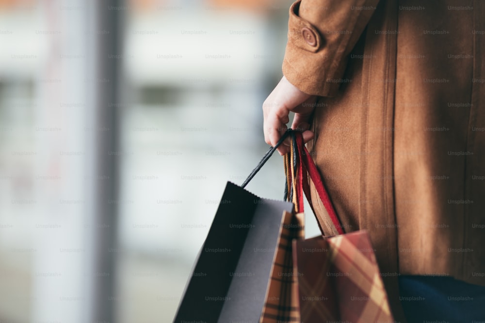 Elegant and modern woman in brown leather jacket or topcoat standing on the city street and holding bunch of shopping bags. Close up shot of woman hand. Lifestyle consumerism theme.