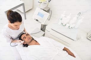 L'estetista professionista sta realizzando una terapia di massaggio facciale elettrico per l'uomo. Il ragazzo sta mentendo con piacere
