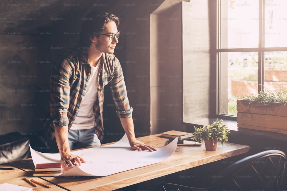 Thoughtful young man in casual wear holding pencil and leaning at the wooden desk with blueprint laying on it