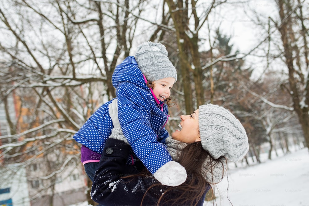 Beautiful young mother with her cute little daughter playing outside in winter nature. Mom lifting girl up.