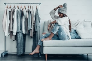 Thoughtful young woman in casual wear adjusting her hat and looking away while sitting on the couch at home near her clothes hanging on the racks