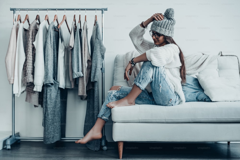 Thoughtful young woman in casual wear adjusting her hat and looking away while sitting on the couch at home near her clothes hanging on the racks