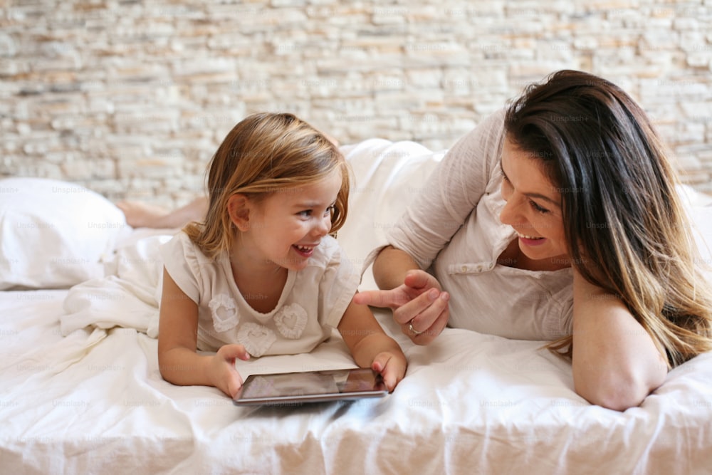 Happy young woman with daughter using digital tablet on bed at home.
