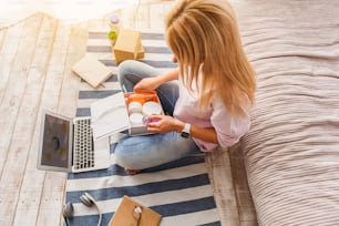 Top view young blond girl buying cosmetic products through internet. She is sitting on floor at home and holding open box. Lady is looking at perfume with interest