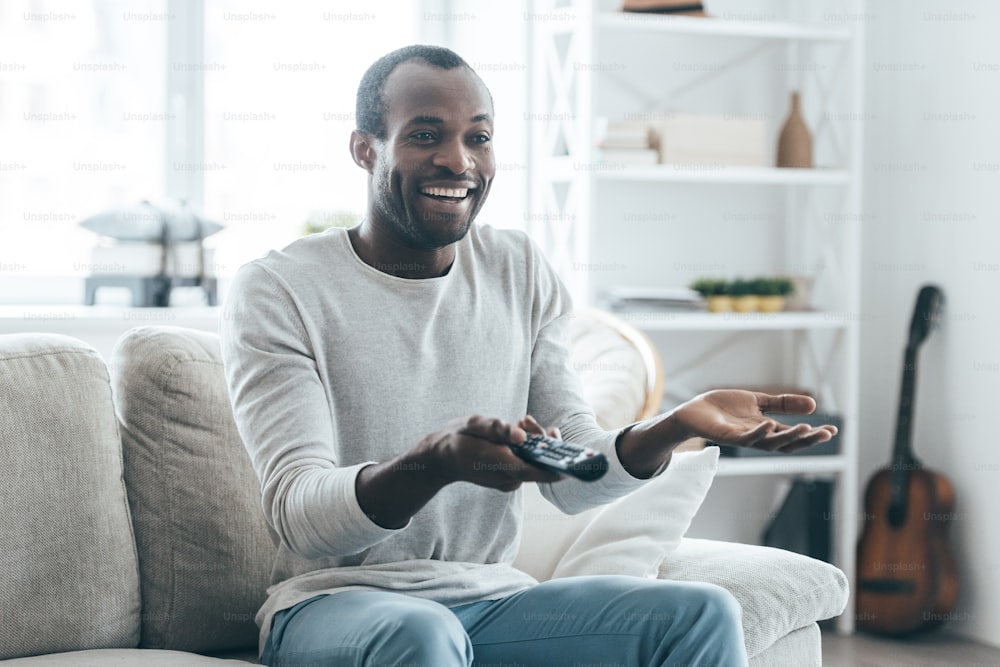 ハンサムな若いアフリカの男性は、自宅のソファに座ってテレビを見て微笑む