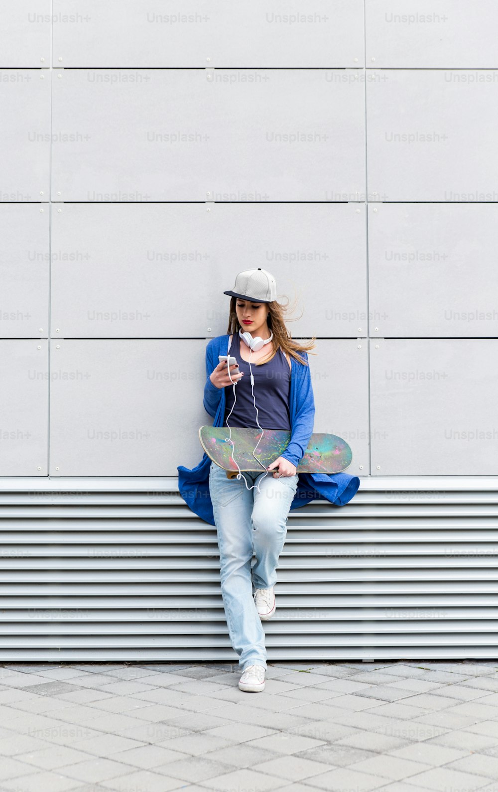 Niña con patineta apoyada en una pared gris moderna, sostiene un teléfono móvil en la mano.