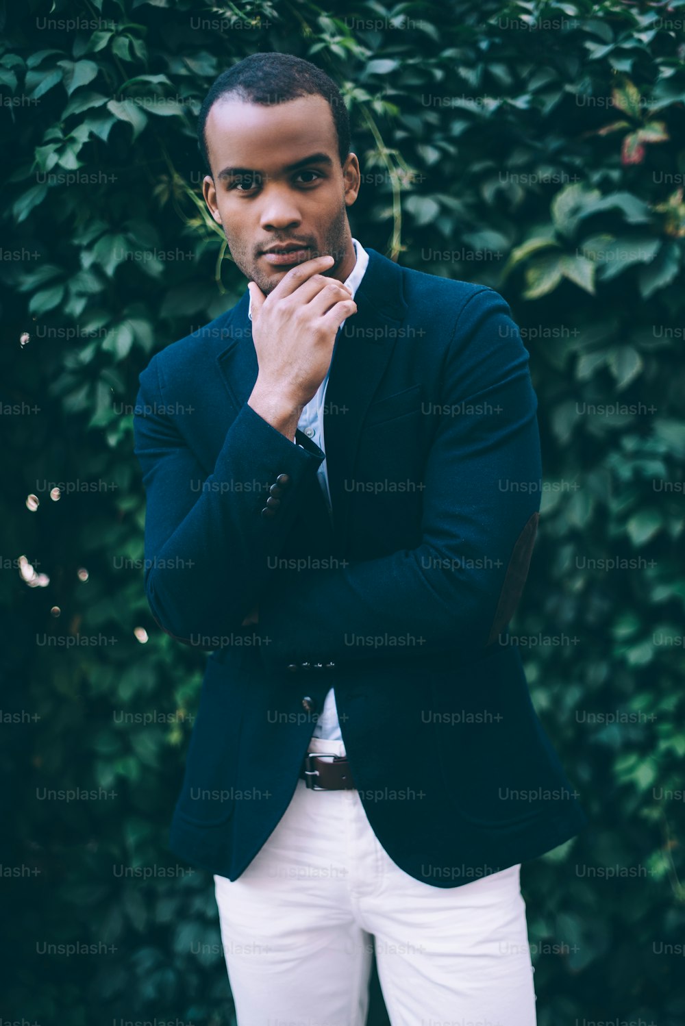 Handsome young African man in smart casual wear holding hand on chin and looking at camera while standing against green plant background outdoors