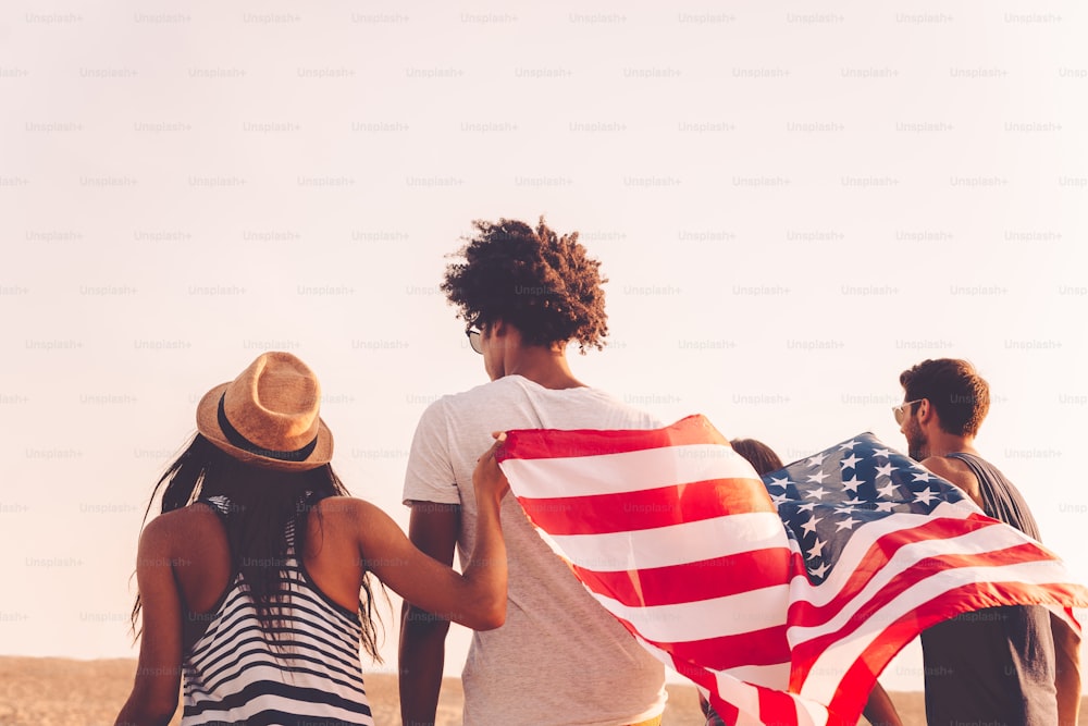 Vista trasera de cuatro jóvenes que llevan la bandera americana mientras corren al aire libre