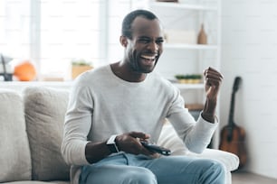 Handsome young African man holding a remote control and laughing while sitting on the sofa at home