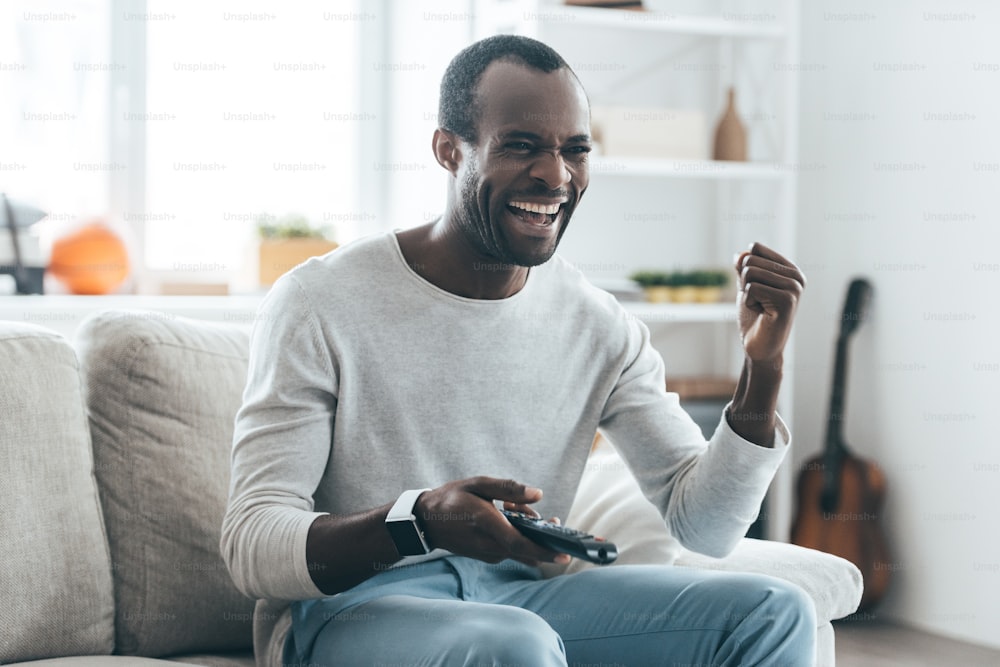 Handsome young African man holding a remote control and laughing while sitting on the sofa at home