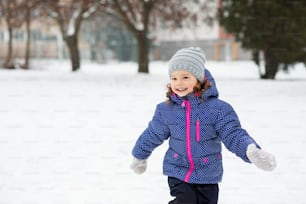 Jolie petite fille en veste bleue et bonnet tricoté jouant dehors dans la nature hivernale, courant partout