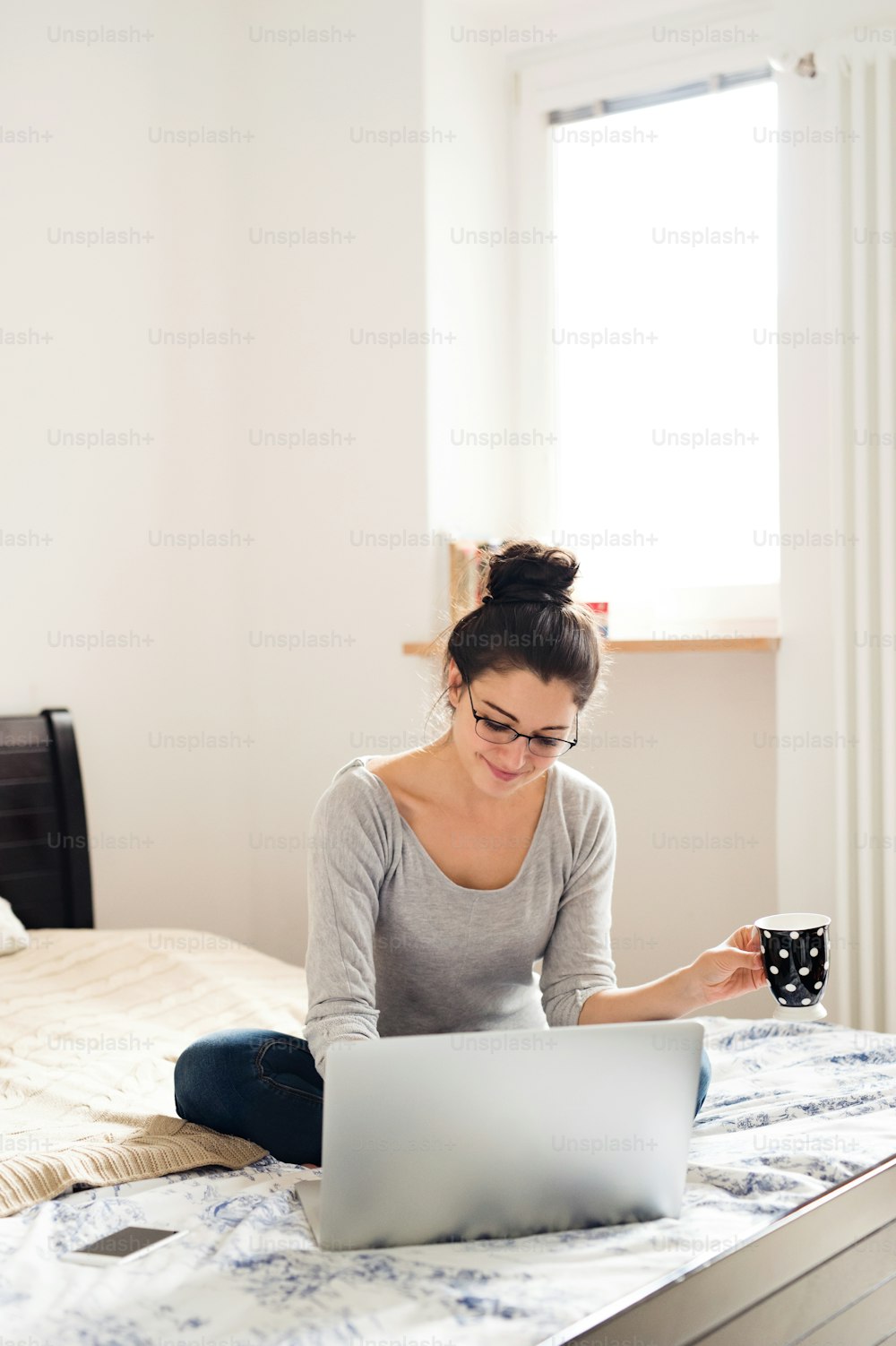 Schöne junge Frau, die auf dem Bett sitzt, am Notebook arbeitet, eine Tasse Kaffee oder Tee in der Hand hält, Home Office.