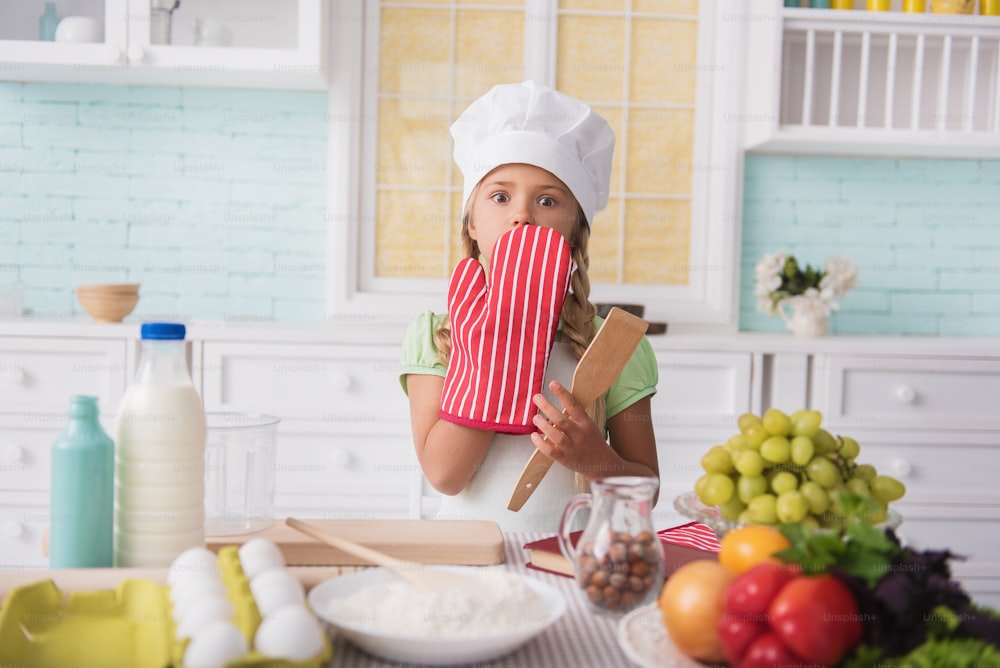 Oops. Cute little girl made mistake in cooking. She is covering mouth with hand in potholder. Child is standing and looking at camera with fear