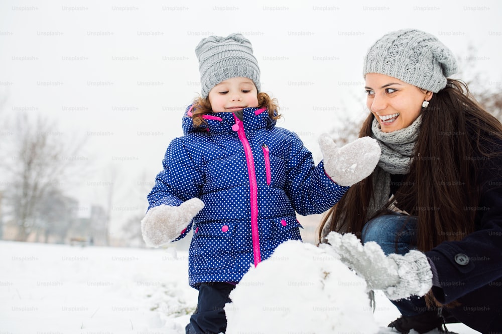 Schöne junge Mutter mit ihrer süßen kleinen Tochter, die einen Schneemann baut und draußen in der winterlichen Natur spielt