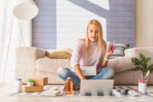 Blond girl is buying cosmetic products in internet with aspiration. She is typing on laptop while holding bottle of perfume. Lady is sitting on floor in her apartment and smiling